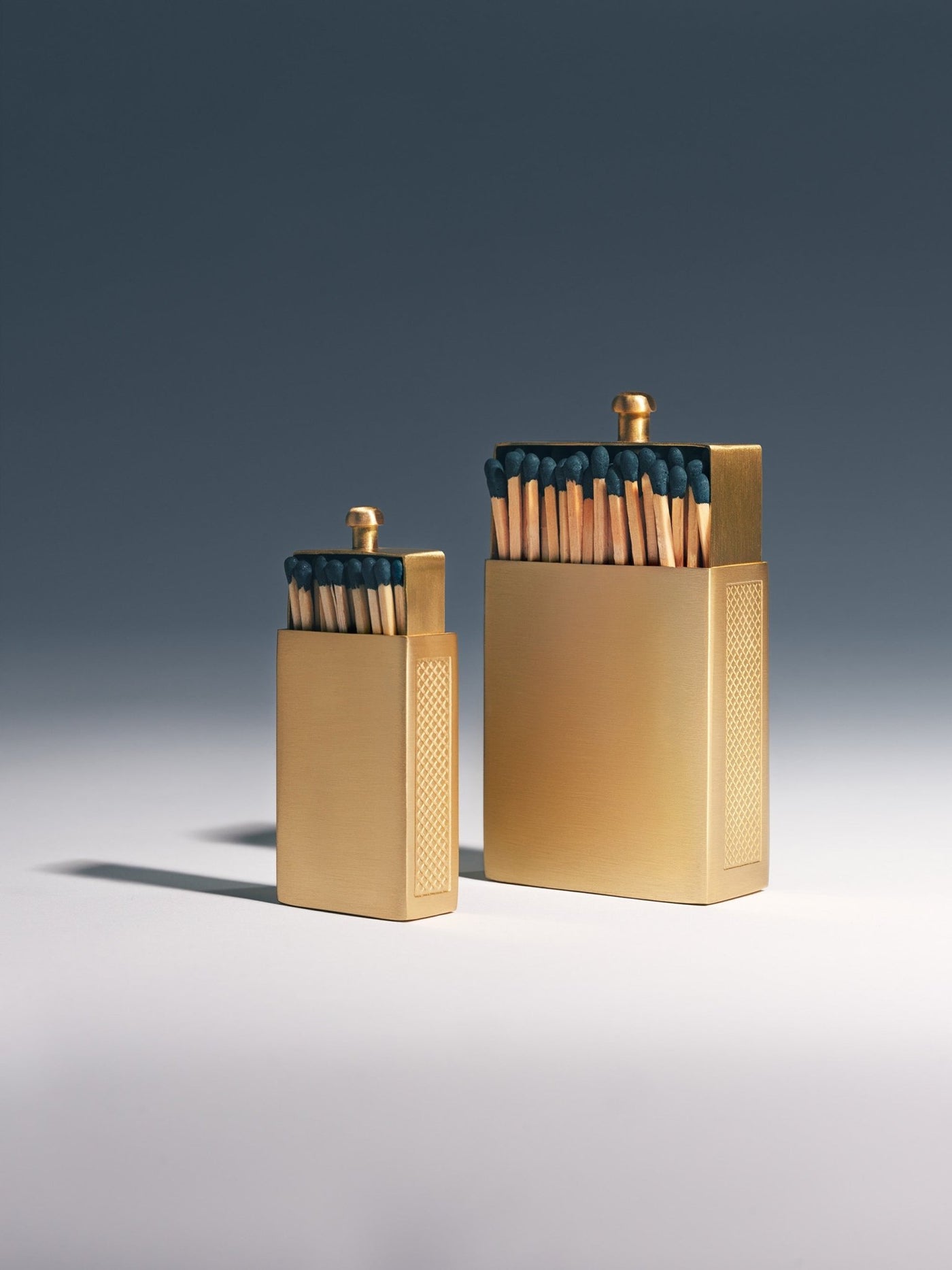Little and Big brushed brass matchbox size comparison, both engraved with "I'm coming to the cottage," studio shot with blue gradient background | Here & Always